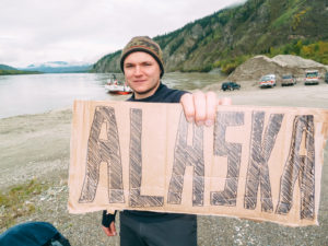 Phillip holder en pappskilt til kameraet, der det står Alaska. I bakgrunnen kan du se fjell og en innsjø.