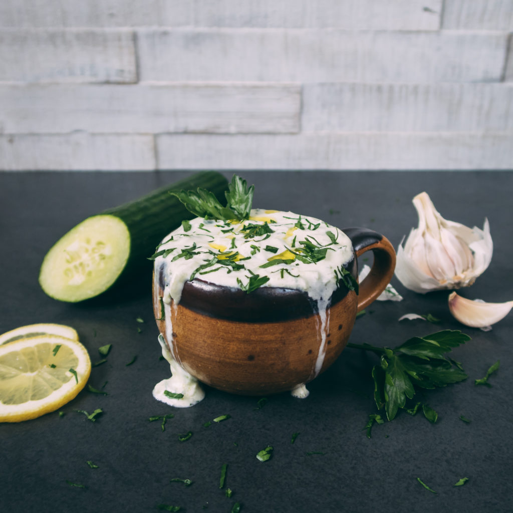 In einer Tasse ist Tzatziki. Daneben liegt ein Stück Gurke, Zitronenscheiben und Knoblauch. Der Tzatziki ist mit Olivenöl und glatter Petersilie verziert.