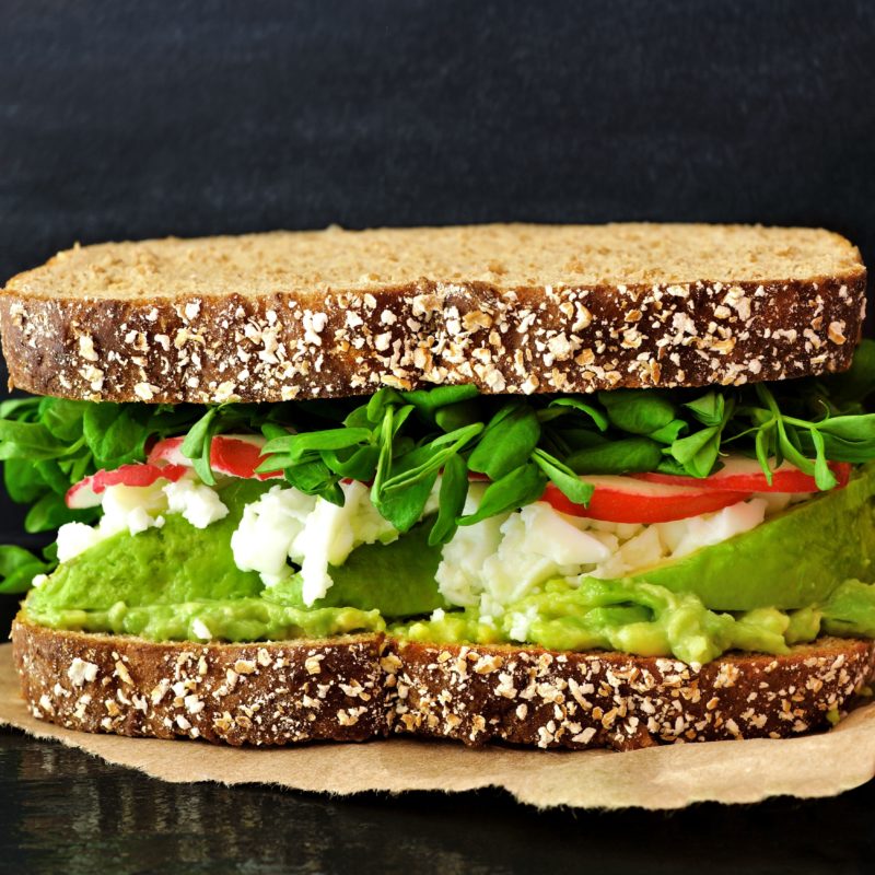 Frontale Sicht auf ein Sandwich mit Avocado, Kresse, Feta und Tomaten. Das Sandwich liegt auf Backpapier.