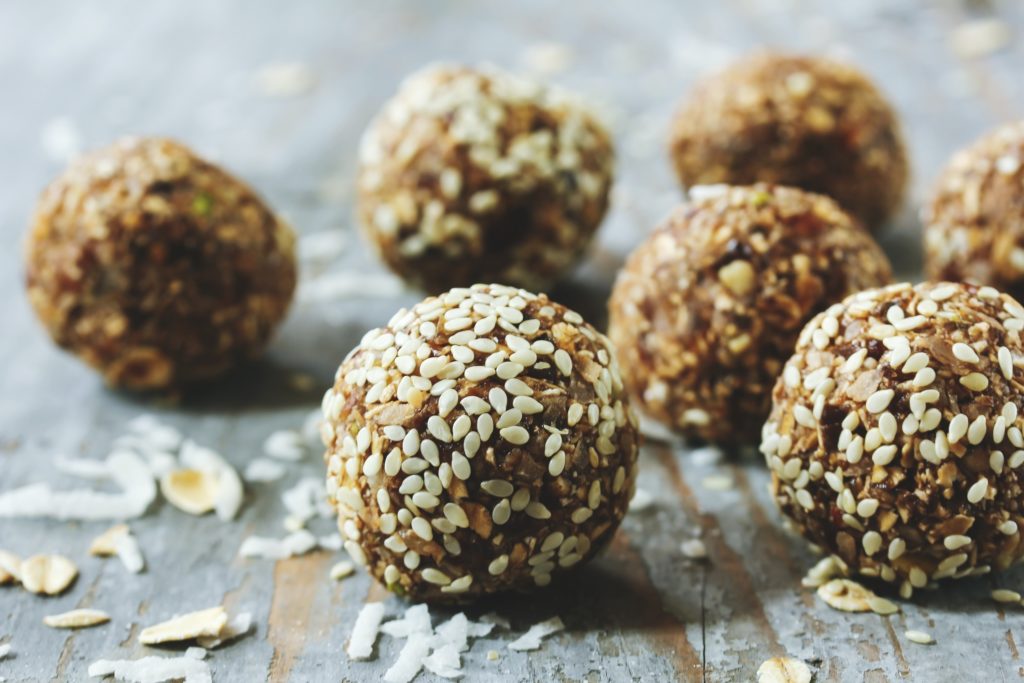 Man sieht mehrere Energy Balls in der Mitte. Die Balls sind mit Sesam ummantelt. Um die Energy Balls herum liegen verstreut Kokosraspeln und Haferflocken.
