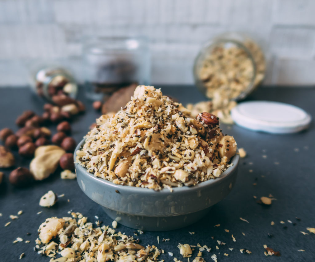 Im Vordergrund sieht man das fertige Paleo Müsli in einer Schüssel. Im Hintergrund liegen vereinzelt Nüsse sowie das Müsli.