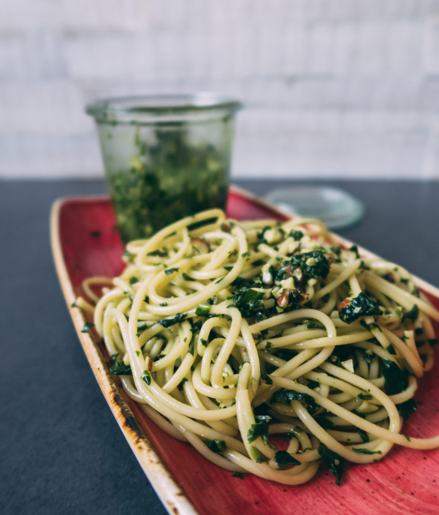 Auf einer roten Schale liegen Spaghetti mit grünem Spinat Pesto. Im Hintergrund steht ein Glas mit Mandel-Spinat-Pesto.