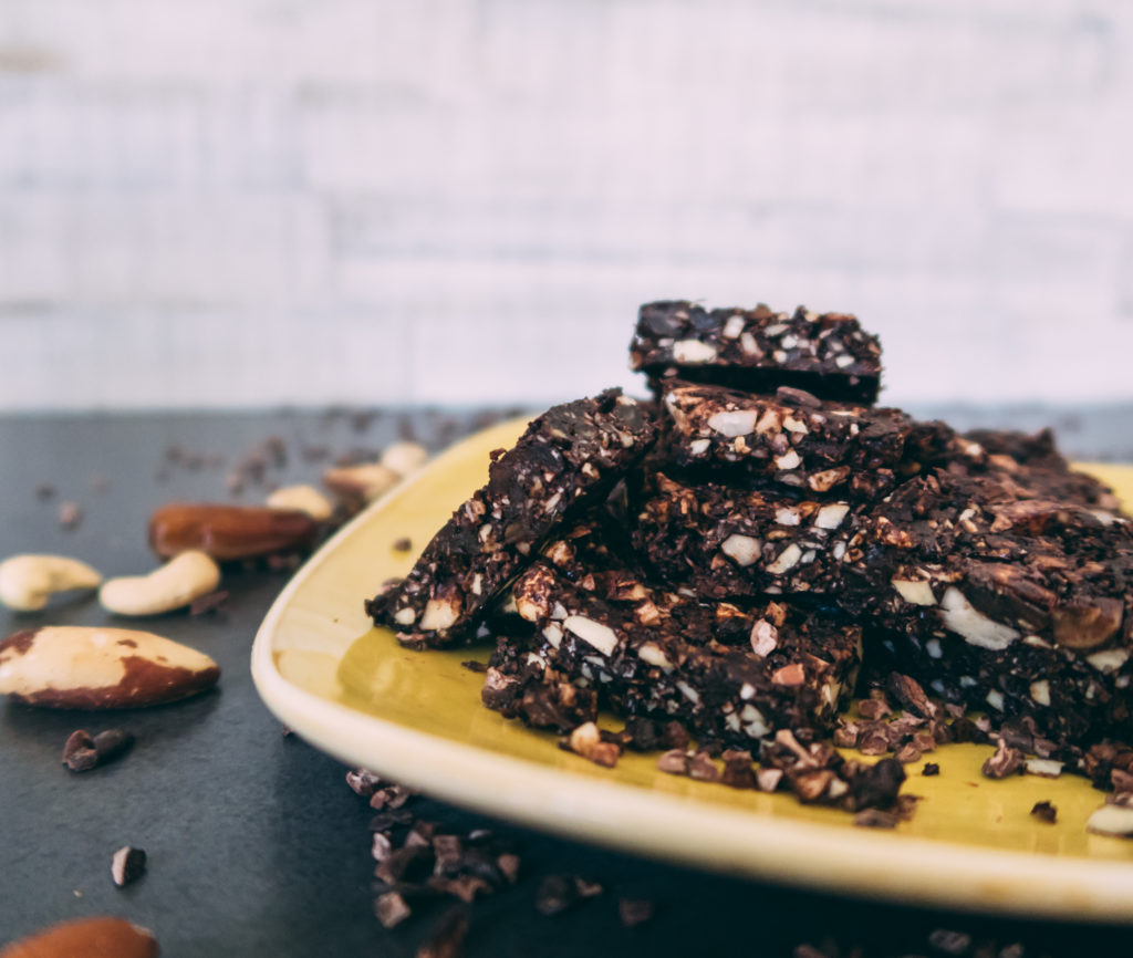 Auf einem gelben Teller von Churchill sind die Raw Brownies drapiert. Um den Teller liegen Paranüsse, Cashewkerne, Datteln sowie Kakaonibs.