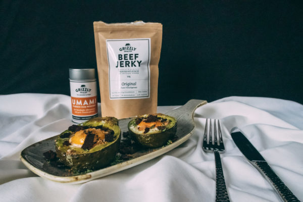 Breakfast Avocado Avocado Frühstück Auf einem Teller von Churchill liegt die Avocado. Im Hintergrund sieht man eine Packung Beef Jerky sowie eine Dose UMAMI-Gewürzmischung.