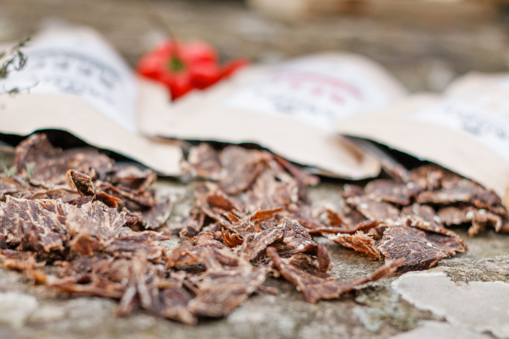 Im Vordergrund sieht man Stücke von Trockenfleisch, im Hintergrund sieht man die offenen Tüten der Sorten Cracked Pepper, Burn Baby Burn und Original
