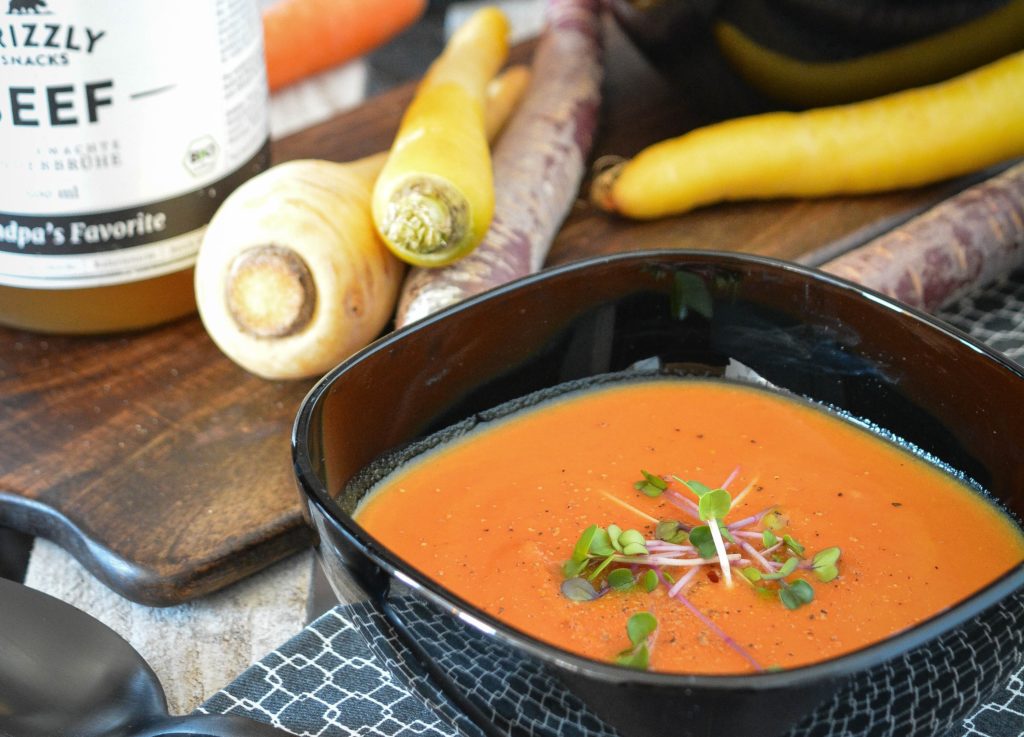 In einer schwarzen Schüssel im Vordergrund ist Rübensuppe zu sehen. Sie ist mit Kresse verziert. Links dahinter steht die Rinder-Kraftbrühe und Grizzlysnacks. Rote Beete, Pastinaken und Möhren liegen zusätzlich im Hintergrund.