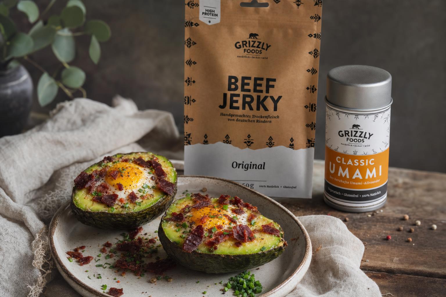 Zwei Avocadohälften mit gebackenem Ei und knusprigen Beef Jerky-Stücken auf Keramikteller, daneben Besteck auf weißem Tuch.