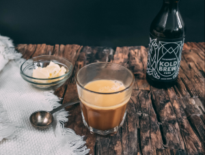 Bulletproof Coffee im Glas, links daneben eine Schale mit etwas Butter und rechts davon eine Flasche Cold Brew von Philosoffee