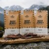 Drei Beutel Beef Jerky liegen nebeneinander auf einem Holzbrett im Freien, im Hintergrund Berge und Natur unscharf erkennbar.