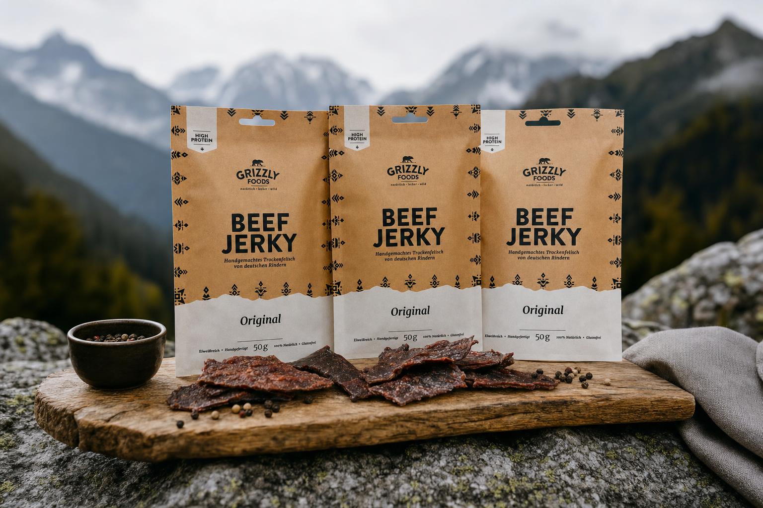 Drei Beutel Beef Jerky liegen nebeneinander auf einem Holzbrett im Freien, im Hintergrund Berge und Natur unscharf erkennbar.