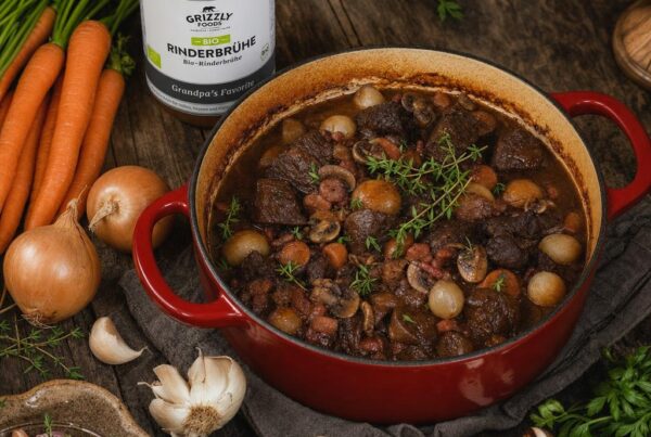 Ein gusseiserner Topf mit Boeuf Bourguignon, garniert mit Kräutern, umgeben von Karotten, Zwiebeln, Knoblauch und Pilzen auf Holztisch.