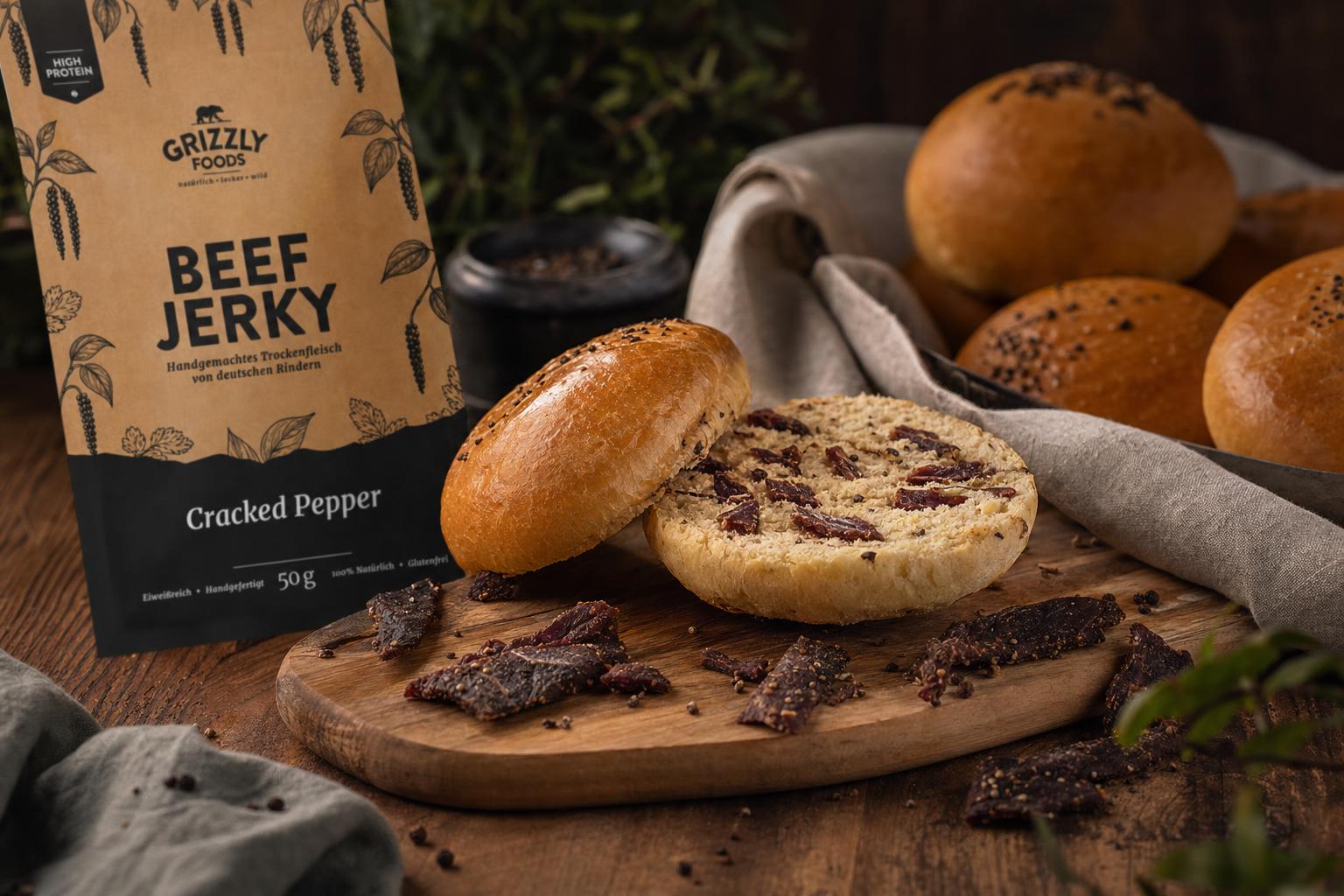 Aufgeschnittenes Burgerbrötchen mit sichtbaren Beef-Jerky-Stücken auf Holzbrett, daneben Jerky-Stücke und weitere Buns im Hintergrund.