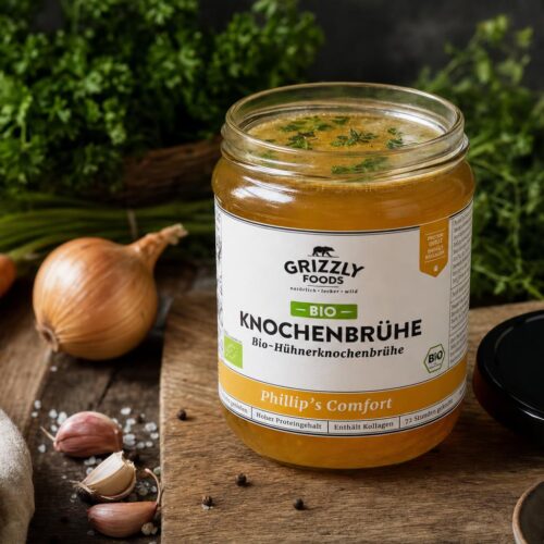 Ein Glas mit goldener Hühnerknochenbrühe, oben frische Kräuter, im Hintergrund Karotten, Zwiebel und Petersilie auf Holz.