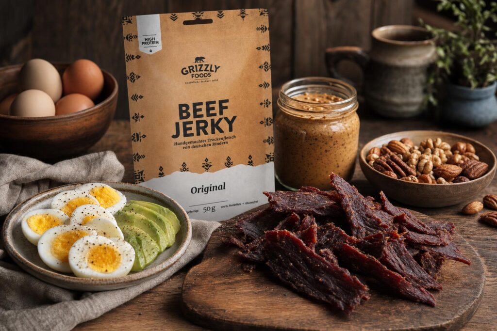 Auf einem Holztisch liegen gekochte Eier mit Avocado, Beef Jerky, gemischte Nüsse, ein Glas Nussmus und ganze Eier im Hintergrund.