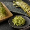 Frisch geriebener Wasabi mit grober, stückiger Textur auf Holzreibe, daneben ein hellgrüner Wasabi-Rhizom.