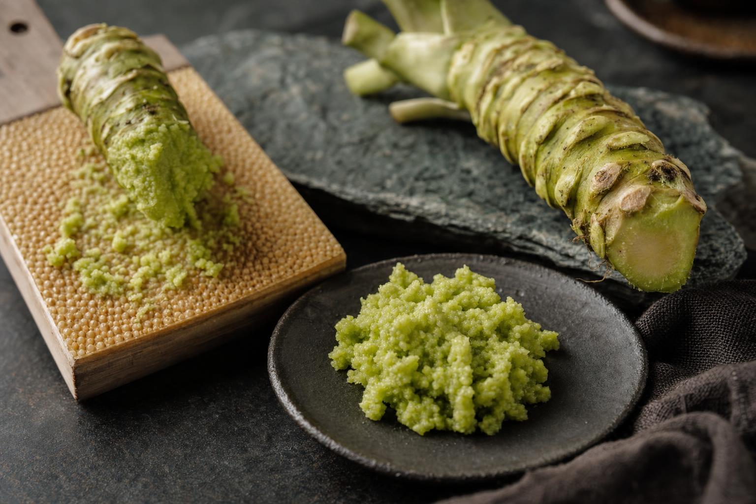 echter Wasabi – Frisch geriebener Wasabi mit grober, stückiger Textur auf Holzreibe, daneben ein hellgrüner Wasabi-Rhizom.