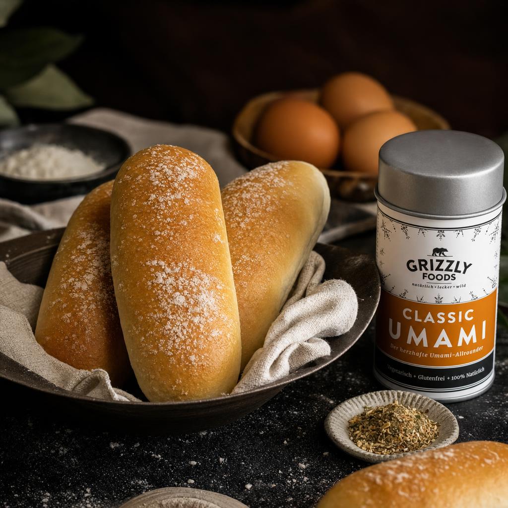 Eine Hand hält ein frisch gebackenes, goldbraunes Hot Dog Brötchen, im Hintergrund weitere Brötchen, Mehl und Eier auf dunkler Fläche.