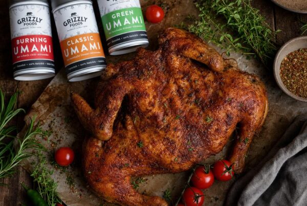 Flach gedrücktes, goldbraun gebratenes Hähnchen auf Backpapier, umgeben von frischen Kräutern, Tomaten und drei Gewürzdosen