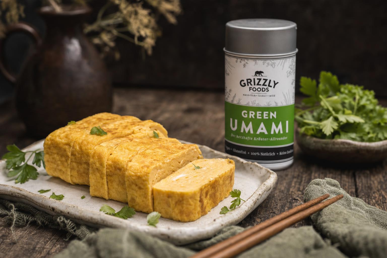 In Scheiben geschnittenes Tamagoyaki auf Keramikteller, garniert mit Petersilie, daneben Holzstäbchen und frische Kräuter auf Holztisch.