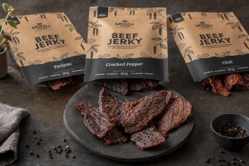 Mehrere Stücke Trockenfleisch liegen auf Stein, im Hintergrund unscharf drei geöffnete Tüten mit verschiedenen Sorten Beef Jerky.