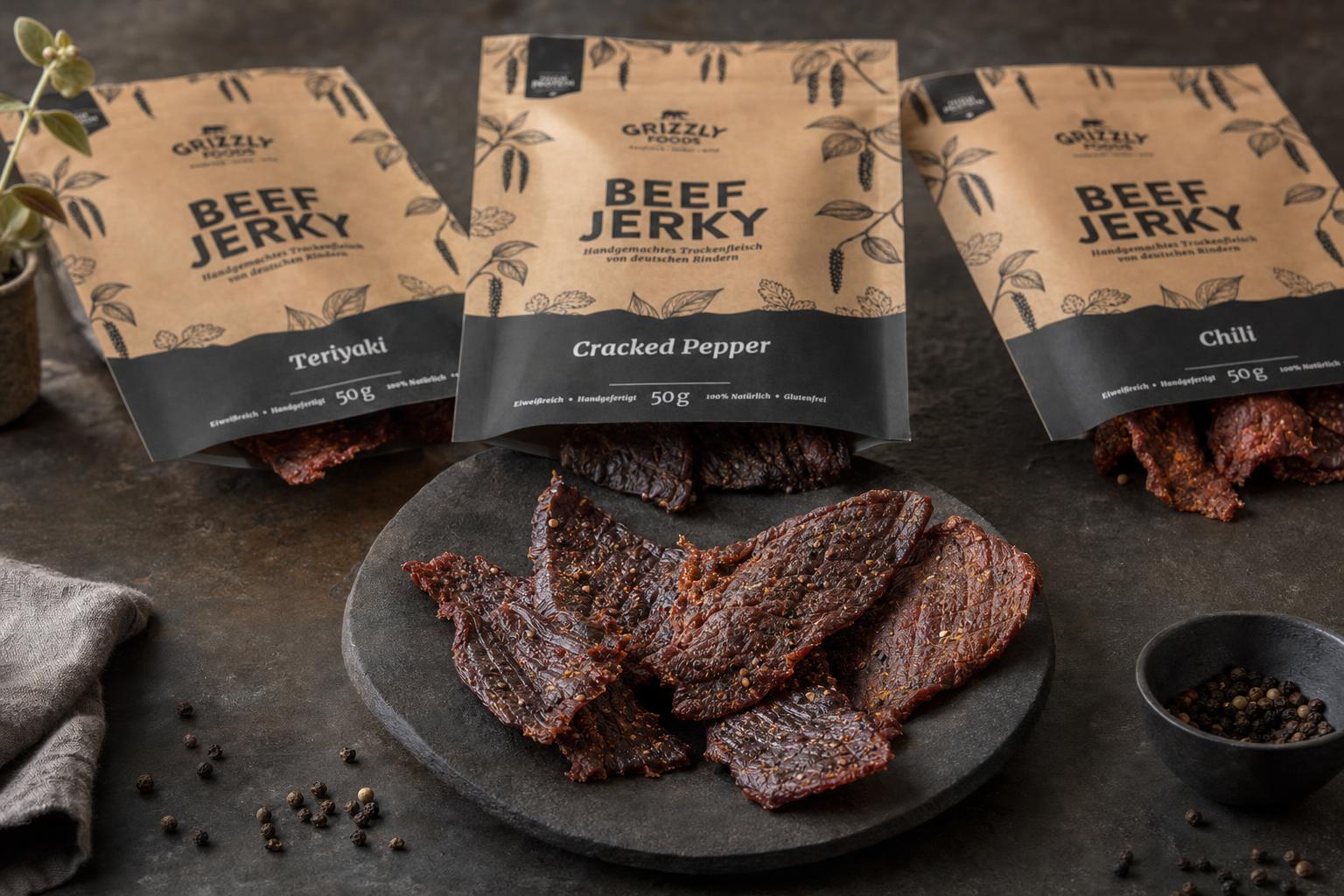 Mehrere Stücke Trockenfleisch liegen auf Stein, im Hintergrund unscharf drei geöffnete Tüten mit verschiedenen Sorten Beef Jerky.