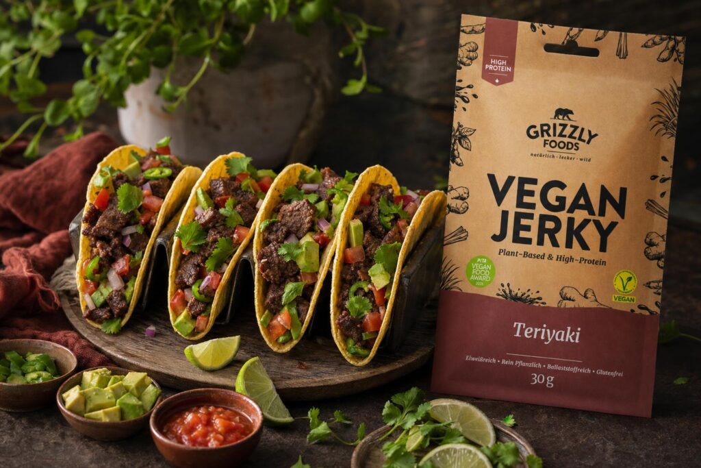 Vier vegane Tacos mit Vegan Jerky, buntem Gemüse und frischen Kräutern auf Holzplatte, dazu Schälchen mit Salsa, Avocado und Limetten.